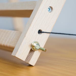 May include: A light-colored wooden frame structure featuring a horizontal bar and a diagonal support beam. The assembly is secured with a metallic wing nut and bolt, and a thin black string passes through a circular hole in the wooden beam.