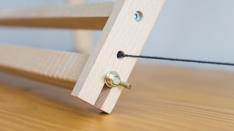 May include: A light-colored wooden frame structure featuring a horizontal bar and a diagonal support beam. The assembly is secured with a metallic wing nut and bolt, and a thin black string passes through a circular hole in the wooden beam.