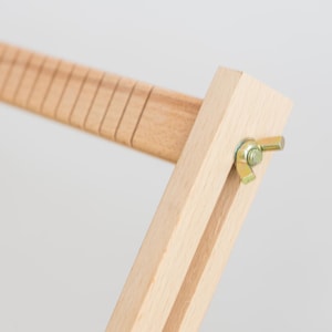 May include: A light-colored wooden frame component featuring a series of evenly spaced vertical grooves along one edge. The piece is connected to a perpendicular wooden arm by a gold-toned metal wing nut and bolt assembly, allowing for adjustment.