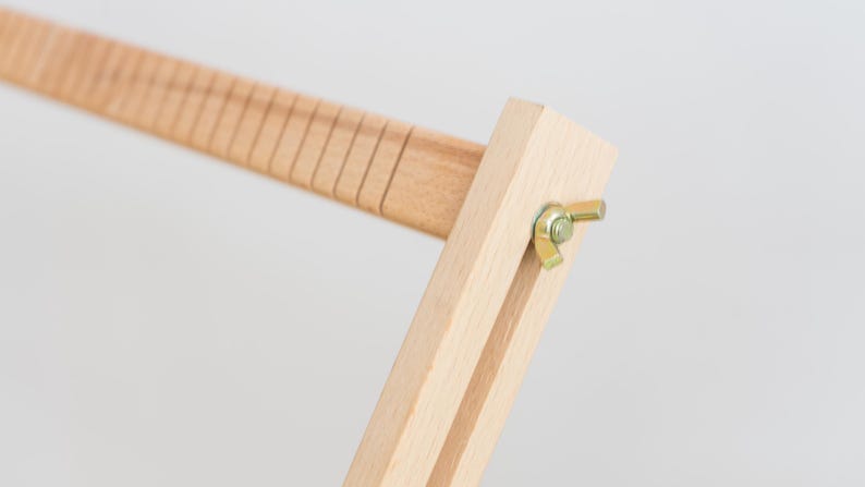May include: A light-colored wooden frame component featuring a series of evenly spaced vertical grooves along one edge. The piece is connected to a perpendicular wooden arm by a gold-toned metal wing nut and bolt assembly, allowing for adjustment.