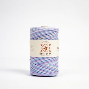 May include: A spool of multi-colored cotton rope in shades of lavender, light blue, and pink. The rope is wrapped around itself, with a paper label wrapped around the middle. The label has text and a cotton flower graphic.