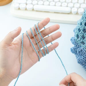 May include: A hand holding light blue yarn, with strands wrapped between the fingers. The yarn appears to be a fine, textured thread. A partially visible crocheted item in shades of blue is in the background, along with a white keyboard.