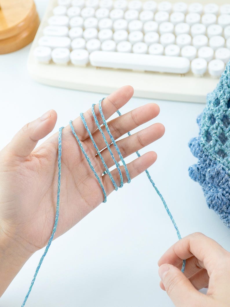 May include: A hand holding light blue yarn, with strands wrapped between the fingers. The yarn appears to be a fine, textured thread. A partially visible crocheted item in shades of blue is in the background, along with a white keyboard.