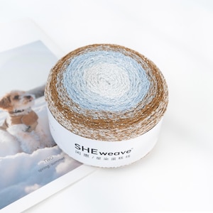 May include: A round ball of yarn with a gradient color scheme, transitioning from brown to light blue and white. The yarn is wrapped around a white cardboard cylinder with the brand name "SHEweave" printed on it. The yarn appears to have a metallic sheen.