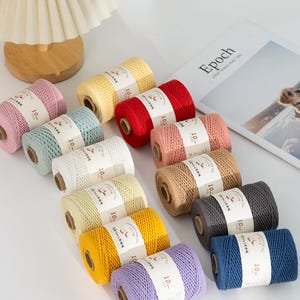 Puede incluir: Una colección de coloridas bobinas de cordón de algodón en varios tonos, incluyendo rosa, amarillo, rojo y azul. Cada bobina tiene una etiqueta blanca con texto. Los cordones están dispuestos sobre una superficie blanca.