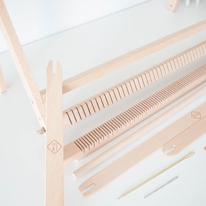 May include: A light-colored wooden weaving loom kit featuring a frame, two slotted heddle bars, two flat shuttle sticks, a small wooden comb, a thin metal needle, and a thin wooden stick, all arranged on a flat surface for assembly and use.