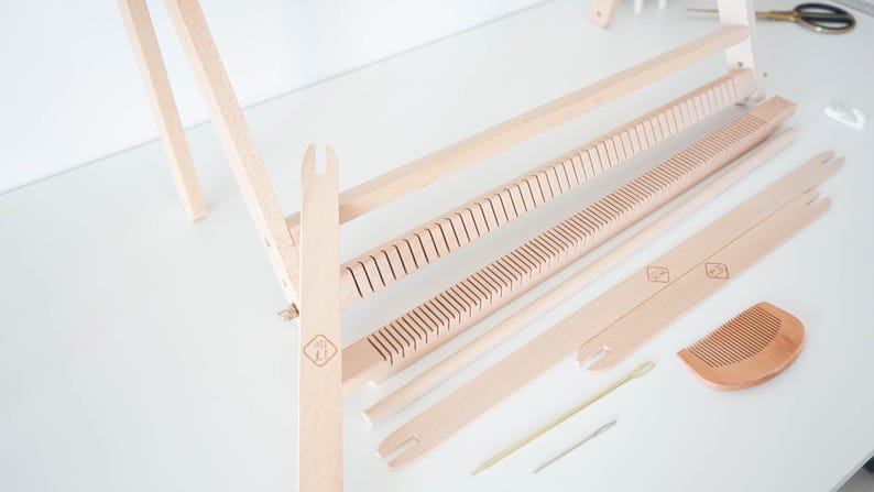 May include: A light-colored wooden weaving loom kit featuring a frame, two slotted heddle bars, two flat shuttle sticks, a small wooden comb, a thin metal needle, and a thin wooden stick, all arranged on a flat surface for assembly and use.