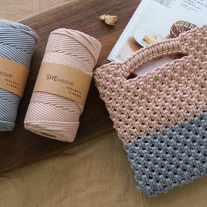May include: A handmade macrame tote bag in beige and gray, with matching spools of cotton cord. The spools are labeled "SHEweave" and are in gray and beige. The bag has a woven design and a handle.