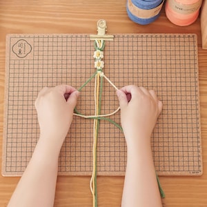 Puede incluir: Primer plano de manos anudando una pulsera con hilos amarillos, verdes y blancos. La pulsera se sujeta con un clip dorado sobre una tabla de corcho cuadriculada. También se ven carretes de hilo y madejas de hilo de bordar.