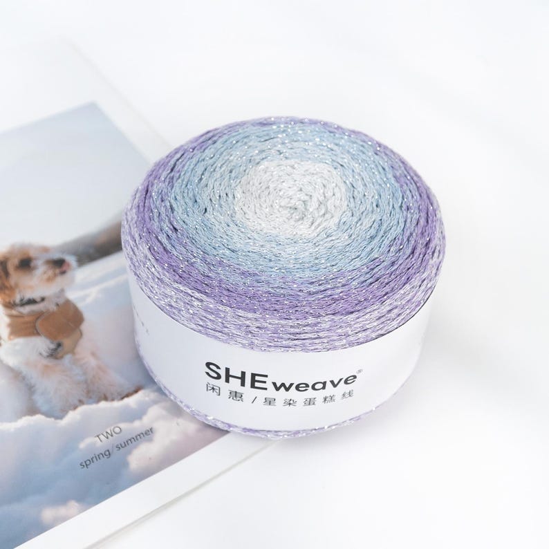 May include: A ball of yarn with a gradient of colors, including white, light blue, and purple. The yarn has a subtle shimmer and is labeled "SHEweave". The image also shows a magazine with a dog on the cover.