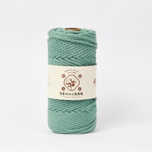 May include: A cylindrical spool of sage green twisted cotton cord wrapped in a cream-colored paper label. The label features a central circular graphic of a cotton plant, brown text, and the characters 4mm, indicating the cord thickness.