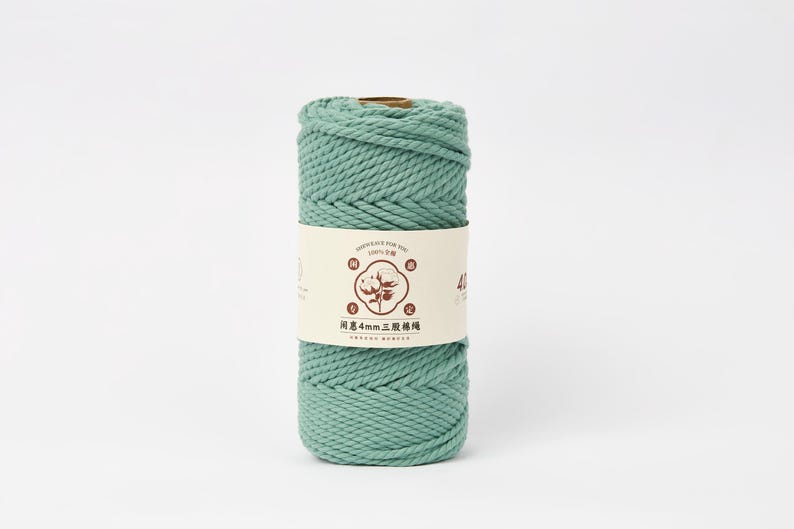 May include: A cylindrical spool of sage green twisted cotton cord wrapped in a cream-colored paper label. The label features a central circular graphic of a cotton plant, brown text, and the characters 4mm, indicating the cord thickness.