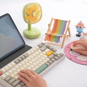 May include: A light green desk fan with yellow blades and a small decorative beach chair with a rainbow-colored woven seat. A keyboard with white and gray keys and a pink mouse pad with a strawberry design are also visible.