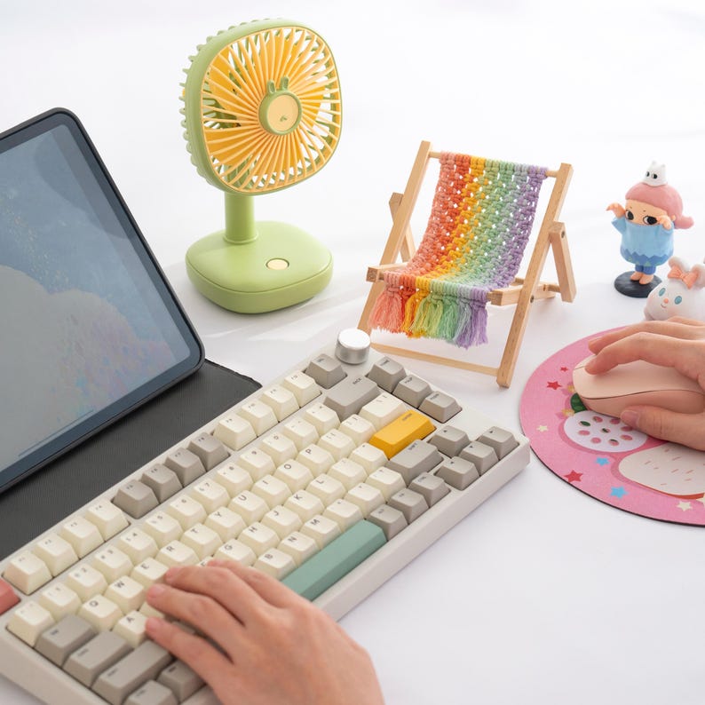 May include: A light green desk fan with yellow blades and a small decorative beach chair with a rainbow-colored woven seat. A keyboard with white and gray keys and a pink mouse pad with a strawberry design are also visible.