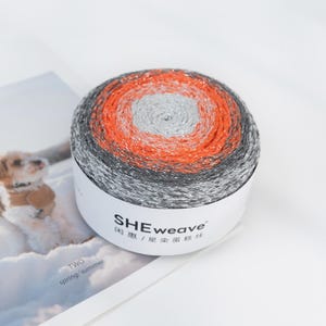 May include: A round skein of yarn with a gradient color scheme. The yarn transitions from silver to orange, then to gray. The label on the yarn reads "SHEweave" in a stylized font. The yarn is likely used for knitting or crocheting projects.