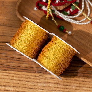Könnte beinhalten: Zwei weiße Kunststoffspulen, die mit leuchtend goldgelbem Garn umwickelt sind. Das Garn hat eine glatte, leicht reflektierende Textur und ist fest um jede Spule gewickelt. Die Spulen sind nebeneinander platziert und zeigen die Farbe.