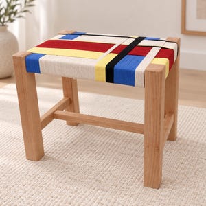 May include: A wooden stool with four square legs and a seat woven from thick fabric straps. The seat features a geometric pattern of intersecting lines and blocks in primary colors including red, yellow, blue, black, and off-white, creating a grid.