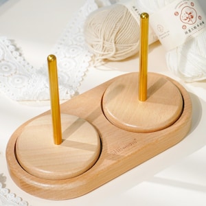 Puede incluir: Un soporte de hilo de madera con dos postes dorados. El soporte tiene un acabado de madera clara y el texto "SHEweave" está impreso en el lateral. Dos ovillos de hilo están en el fondo.