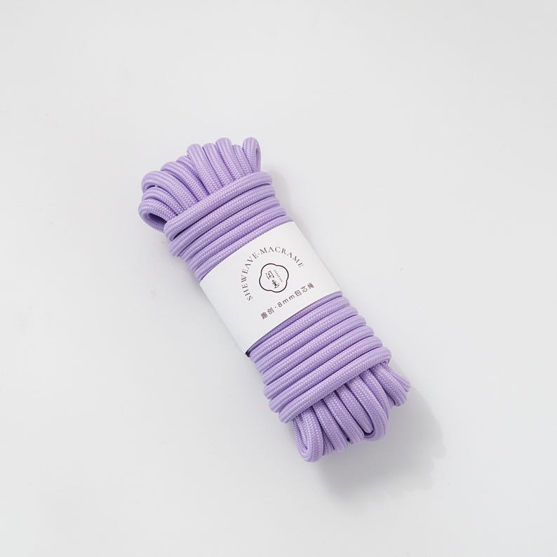 May include: A bundle of lavender macrame cord with a white label wrapped around it. The label reads "SHEWEAVE-MACRAME" and "8mm". The cord is tightly wound and ready for crafting projects. The background is a solid white.