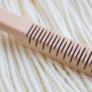 May include: A light-colored wooden weaving tool with a rectangular handle and a series of evenly spaced vertical slots along one side, positioned diagonally across a background of thick, cream-colored twisted yarn strands.