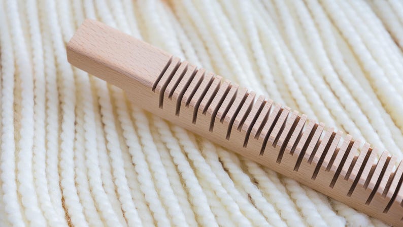 May include: A light-colored wooden weaving tool with a rectangular handle and a series of evenly spaced vertical slots along one side, positioned diagonally across a background of thick, cream-colored twisted yarn strands.