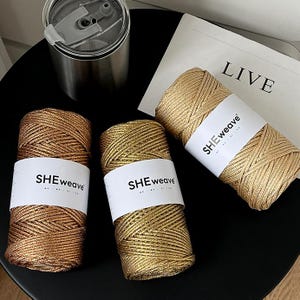 Puede incluir: Tres carretes de hilo dorado metálico con etiquetas blancas que dicen "SHEweave". Los carretes están colocados sobre una superficie negra, junto con un vaso de acero inoxidable y un papel con la palabra "LIVE". El hilo está bien enrollado.