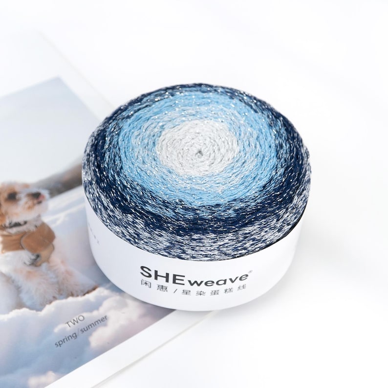 May include: A round skein of yarn in shades of blue, white, and silver. The yarn is wound in concentric circles, creating a gradient effect. The label reads "SHEweave" in white text, with additional text in Chinese characters.