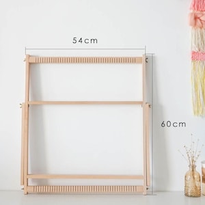 May include: A light-colored wooden weaving loom featuring a rectangular frame with notched top and bottom bars. The frame includes side supports with wing nuts and measures 54 cm in width and 60 cm in height as indicated by the printed text.