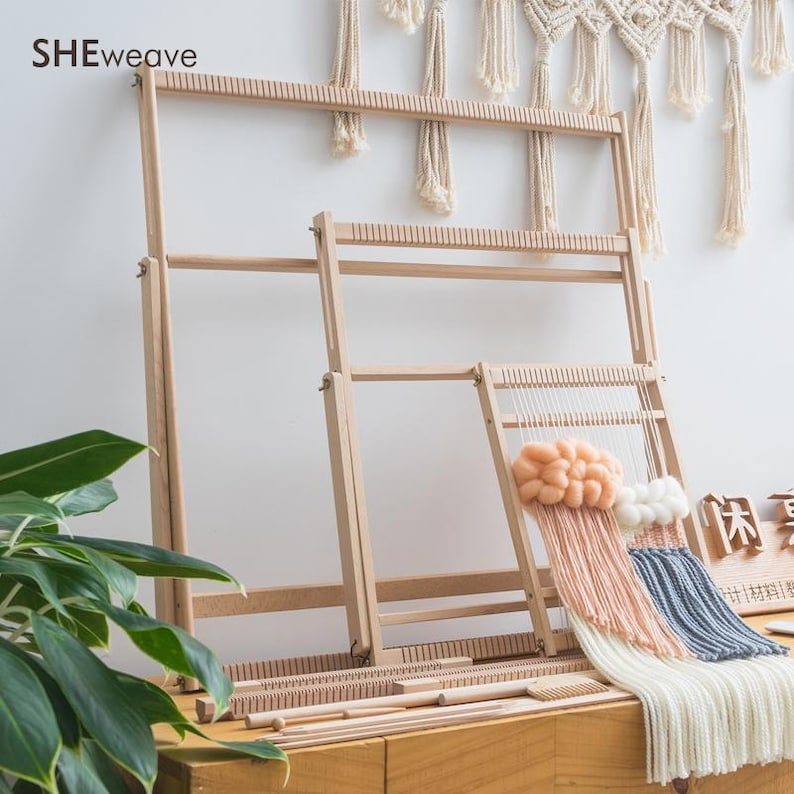 May include: Three light-colored wooden weaving looms of varying sizes arranged in a row. The smallest loom features a partially completed woven tapestry with peach and white yarn clouds and long, hanging fringe in white, peach, and blue.