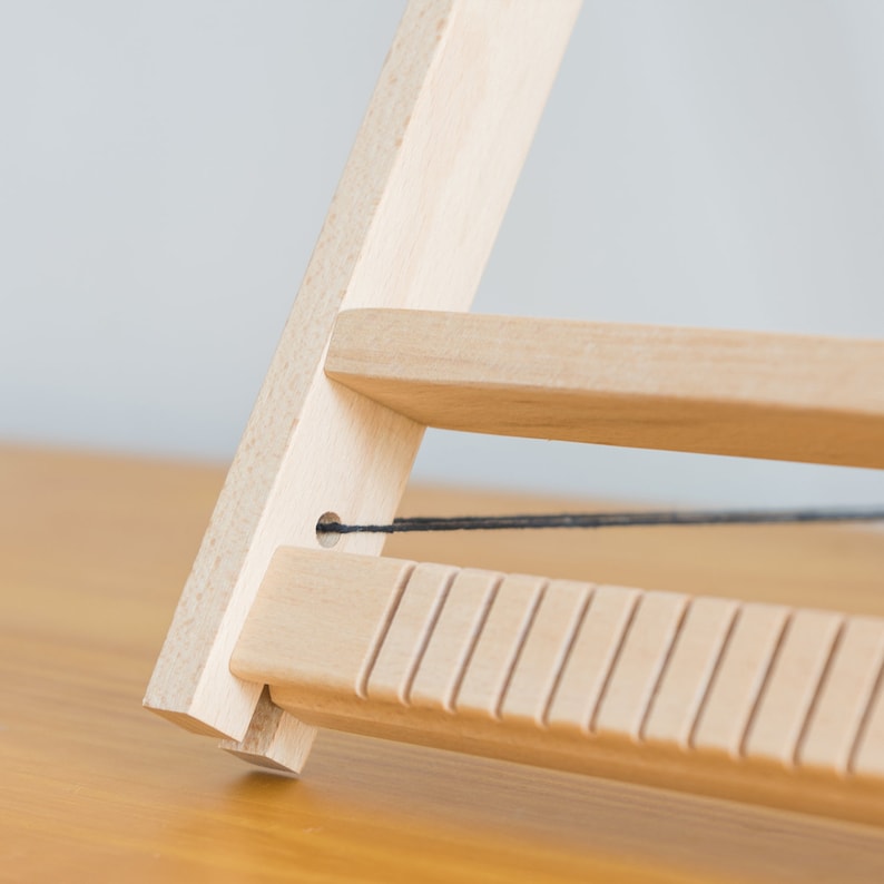 May include: Close-up of a wooden weaving loom, featuring a light-colored wood construction. The loom has a triangular frame with horizontal bars and a series of vertical grooves. A black thread is strung across the frame, indicating its use for weaving.
