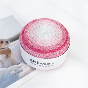 May include: A ball of yarn with a gradient of colors, transitioning from white to pink and then to a deeper magenta. The yarn is in a white container with the text "SHEweave" and Chinese characters. The image is on a white surface.