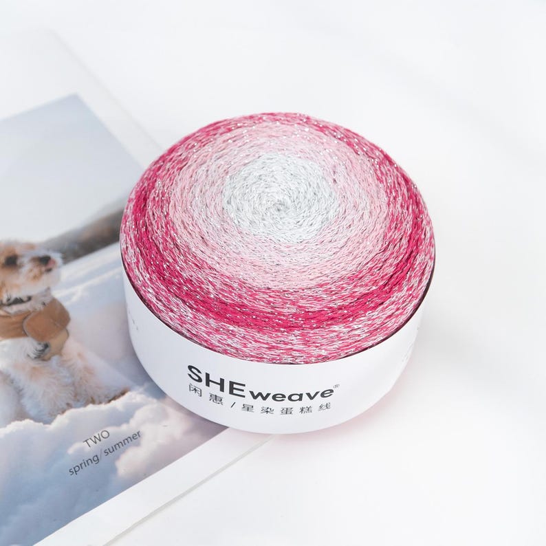 May include: A ball of yarn with a gradient of colors, transitioning from white to pink and then to a deeper magenta. The yarn is in a white container with the text "SHEweave" and Chinese characters. The image is on a white surface.