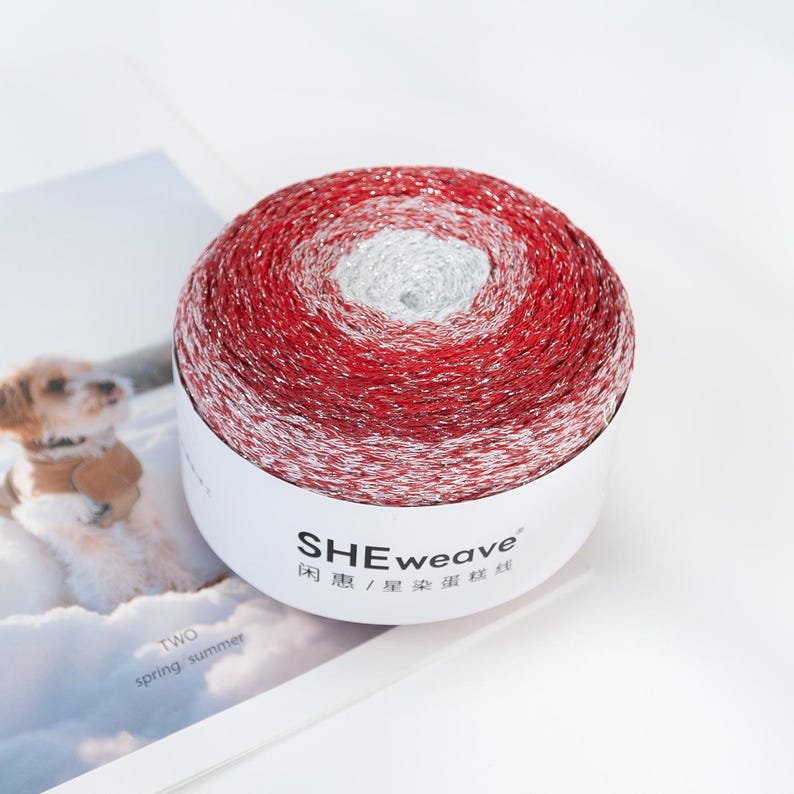 May include: A ball of red, white, and silver yarn with the text "SHEweave" on the label. The yarn is wound in concentric circles, creating a gradient effect. The image also includes the text "TWO spring/summer".