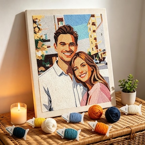 May include: A rectangular canvas featuring a textured needlepoint portrait of a smiling couple standing together. The artwork displays a man in a white shirt and a woman in a pink top against a city background. Several small yarn bobbins in various colors are arranged in front of the canvas.