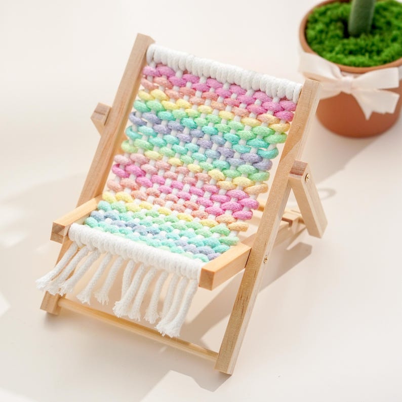 May include: A miniature wooden deck chair with a rainbow-colored woven seat. The chair has a natural wood frame and white fringe along the bottom. The seat features pastel shades of pink, yellow, green, and blue. A small decorative item.