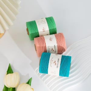 Puede incluir: Tres carretes de hilo de colores verde, coral y turquesa. Cada carrete está envuelto con una etiqueta blanca que incluye texto. El hilo tiene 10 metros de largo. Los carretes están colocados sobre una superficie blanca.