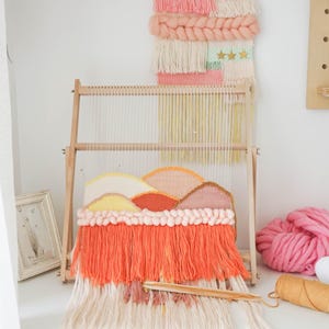May include: A wooden weaving loom holding a partially completed tapestry with a mountain landscape design in shades of peach, coral, and cream. The loom features vertical warp threads and a wooden shuttle resting on the long, vibrant coral fringe.