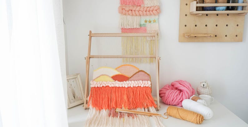 May include: A wooden weaving loom holding a partially completed tapestry with a mountain landscape design in shades of peach, coral, and cream. The loom features vertical warp threads and a wooden shuttle resting on the long, vibrant coral fringe.