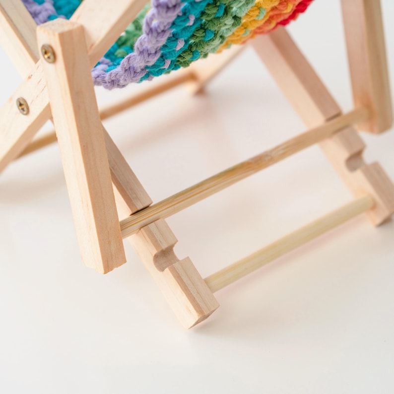 May include: A miniature wooden deck chair with a rainbow-colored crocheted seat. The chair has a natural wood finish and is designed with a folding frame. The seat features a vibrant pattern of red, orange, yellow, green, blue, and purple.