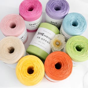 Op de afbeelding: Een verzameling van acht cilindrische klossen raffiagaren in diverse kleuren, waaronder roze, lavendel, lichtblauw, limoengroen, geel, oranje en beige. Elke klos is voorzien van een wit papieren label met de tekst SHEweave en 150g / 300m.