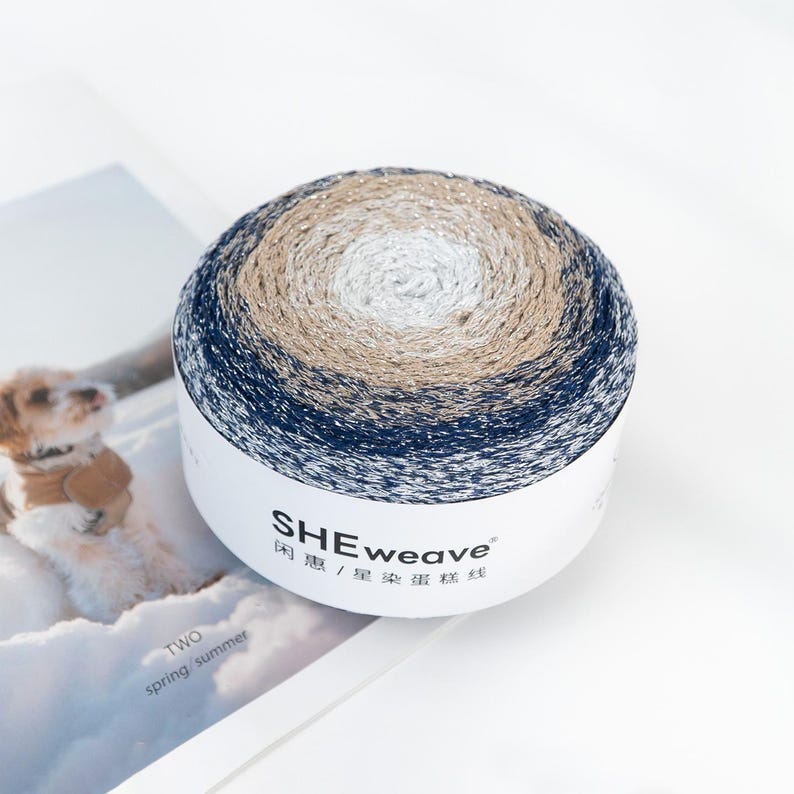 May include: A round skein of yarn in a white container. The yarn is a gradient of colors, including navy blue, tan, white, and gold. The container has the text "SHEweave" printed on it. The yarn is sitting on a magazine.