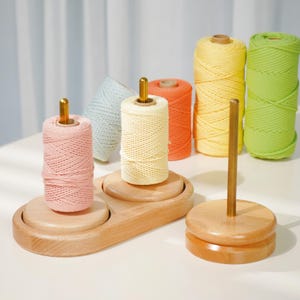 May include: A wooden thread spool holder with two spools of pink and cream-colored thread. Additional spools of thread in yellow, green, orange, and light blue are in the background. The holder has a gold-colored metal rod.