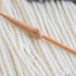May include: A light-colored wooden knitting needle with a tapered point and a rounded knob at the opposite end. The needle features a small, square-shaped logo stamped onto the thicker section of the shaft, showcasing a natural wood grain finish.