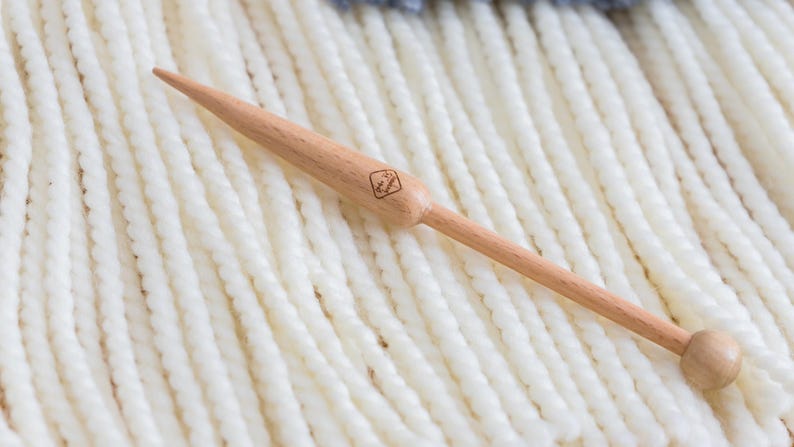 May include: A light-colored wooden knitting needle with a tapered point and a rounded knob at the opposite end. The needle features a small, square-shaped logo stamped onto the thicker section of the shaft, showcasing a natural wood grain finish.