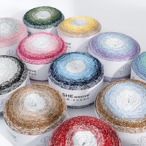 Puede incluir: Una colección de madejas de hilo en varios colores, incluyendo rosa, azul y dorado. Cada madeja está enrollada en forma circular y se asienta sobre una base de cartón blanco. El texto "SHEweave" es visible en algunas bases.