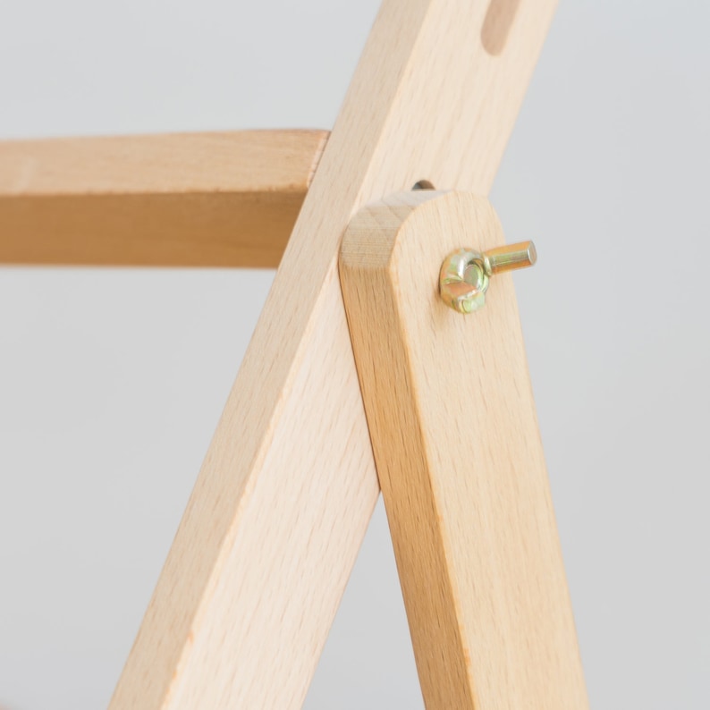 May include: Close-up of a wooden chair joint. The chair is made of light-colored wood, with a visible bolt and nut connecting two wooden pieces. The chair appears to be new and unused, with a simple, clean design.