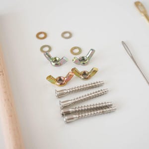May include: A hardware assembly kit containing two light-colored wooden dowels, four metal washers, four metal wing nuts, four threaded metal screws, one silver-toned sewing needle, and one long, thin, gold-toned metal rod with an eyelet at one end.