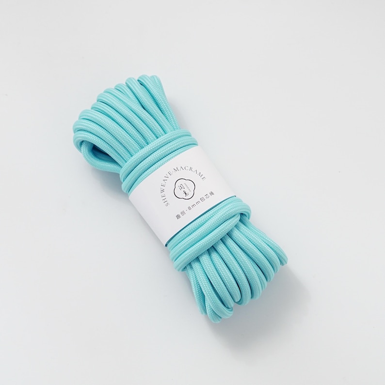 May include: A bundle of light blue macrame cord. The cord is wrapped with a white label that has black text and a logo. The cord appears to be 8mm in diameter. The cord is tightly wound and ready for crafting.