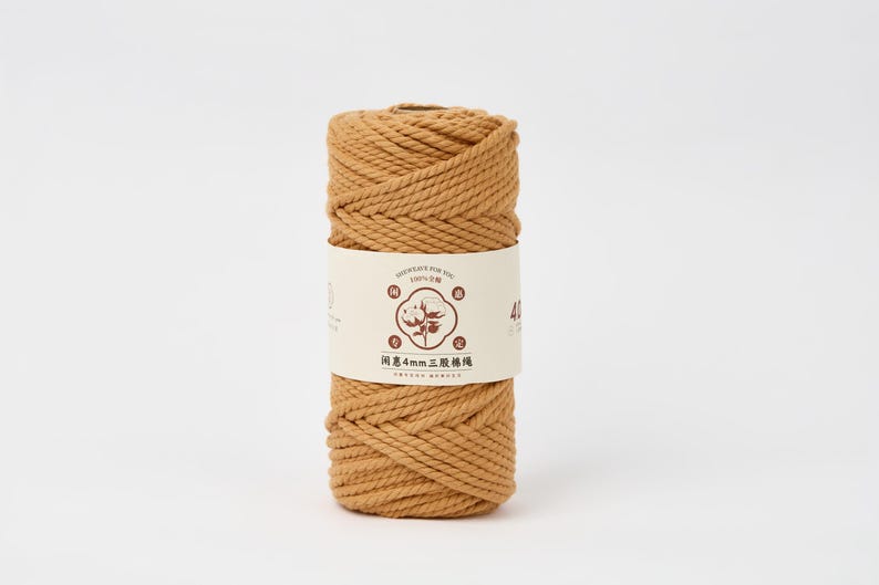 May include: A cylindrical spool of golden-brown twisted cotton cord wrapped in a cream-colored paper label. The label features a central circular graphic of a cotton plant, Chinese characters, and the text 4mm, indicating the cord thickness.