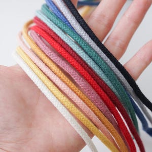 Puede incluir: Una mano sostiene una colección de cordones coloridos. Los cordones son de varios colores, incluyendo blanco, amarillo, rosa, rojo, verde azulado, gris y negro. Los cordones están tejidos apretados y parecen estar hechos de un material duradero.